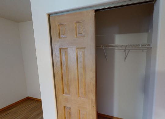 A wooden sliding closet door is partially open, revealing an empty closet with a white wire shelf. The room has light-colored walls and wood flooring. Amber Ridge Apartments.