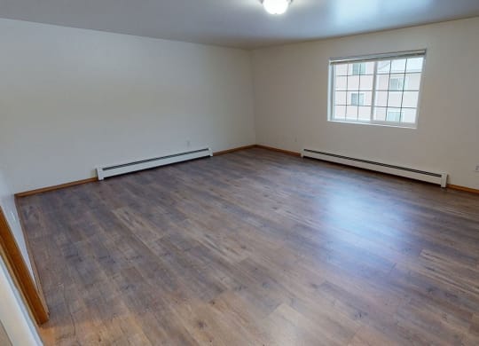 Empty bedroom with light wood flooring, white walls, and a large window letting in natural light. A ceiling light fixture is visible. Cozy and spacious. Amber Ridge Apartments.