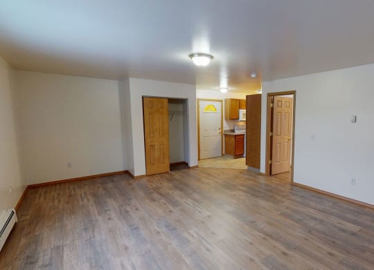 Spacious empty livingroom with wooden floors, white walls, and two open wooden doors leading to a kitchen. A closet with sliding doors is visible. Calm ambiance. Amber Ridge Apartments.