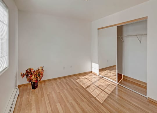 Minimalist empty bedroom with hardwood floor and white walls. A large window casts sunlight, highlighting a flower arrangement in a vase. Peaceful ambiance. Amber Ridge Apartments.