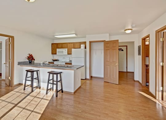 Spacious kitchen with light wood cabinets, white appliances, and a breakfast bar with two stools. Sunlight streams in, casting shadows on hardwood floor. Cozy and inviting. Amber Ridge Apartments.