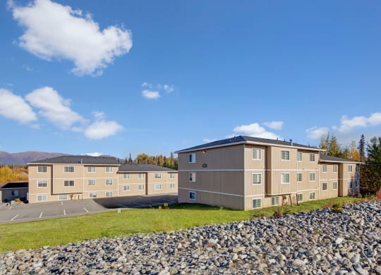 Three beige apartment buildings under a clear blue sky, surrounded by trees with autumn foliage and mountains in the distance, convey tranquility. Amber Ridge Apartments.