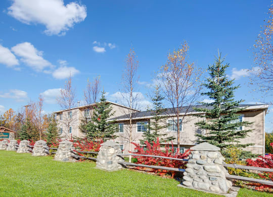 Stone pillars and wooden fence enclose a building surrounded by vibrant fall trees under a bright blue sky, conveying a serene, autumnal atmosphere. Amber Ridge Apartments.