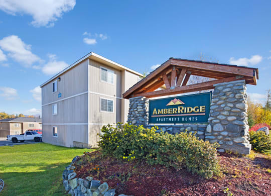 Sign for "Amber Ridge Apartment Homes" on a stone structure under a clear blue sky. Nearby are beige apartment buildings and a parked truck.