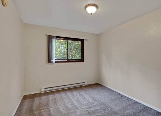 Empty bedroom with beige walls, gray carpet, and a small window with white blinds. Sunlight filters through leaves outside, creating a calm atmosphere. Arbor Ridge Apartments.