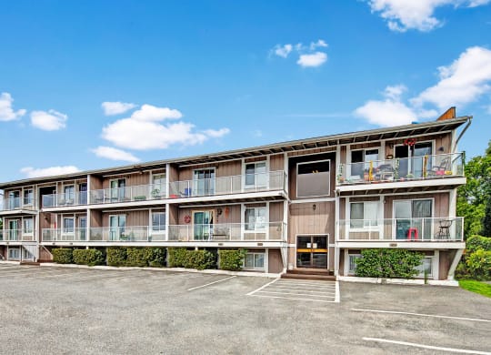 Three-story beige apartment building with balconies and potted plants. An empty parking lot is in front, under a clear blue sky with scattered clouds. Arbor Pointe Apartments.