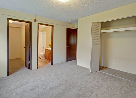 Empty bedroom with beige carpet, open closet with white doors on the right, and two wooden doors on the left leading to a bathroom and another room. Arctic Sun Apartments.