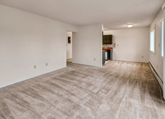 A spacious, neutral-toned living room with beige carpeting, white walls, and an open layout leading to a kitchen. Natural light filters through large windows. Arctic Sun Apartments.
