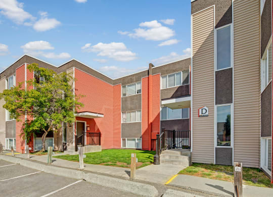Arctic Sun Apartments building with red and beige siding, two stories, and a "D" sign for units 288-350. Trees and parking spaces in the foreground. Sky is clear with scattered clouds.