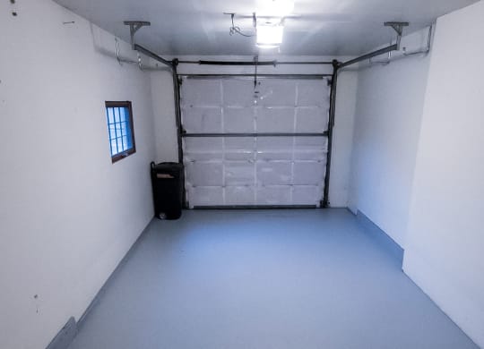 Empty garage here at Birchwood Apartments with white walls and light gray floor, bright overhead light, closed garage door at the back, and a small window on the left. Minimalistic and clean.