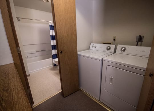 Compact laundry room here at Birchwood Apartments with a washer and dryer next to a bathroom featuring a tub. A blue and white striped shower curtain adds a pop of color.