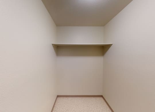 Empty beige closet here at Birchwood Homes Apartments with a single overhead shelf, beige walls, and a textured beige floor. The space conveys simplicity and potential for organization.