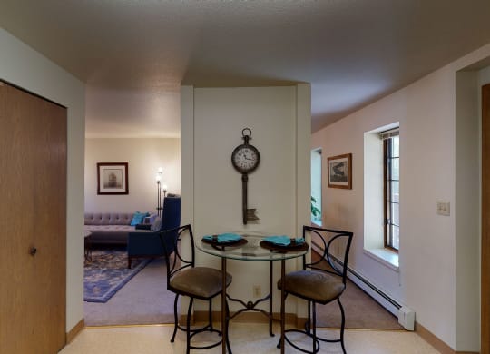 Cozy room here at Birchwood Apartments with a small dining nook featuring a round glass table and two chairs. A large key-shaped clock hangs on the wall. A living area with a sofa and framed art is visible in the background.