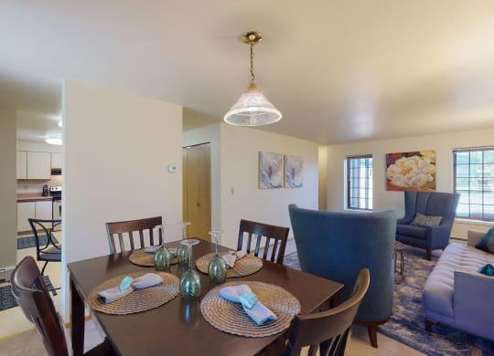 Dining room view of a cozy home with a dark wood table set for four, elegant place settings, and a bright living area featuring plush blue chairs and large windows. Birchwood Homes Apartments.