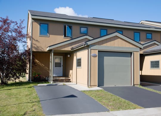 Two-story beige townhouse with a one-car garage, white front door, and small porch. Manicured lawn and a tree under a clear blue sky. Calm suburban setting. Birchwood Homes.