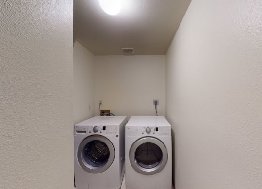 Compact laundry room here at Birchwood Homes Apartments with white walls, featuring a front-loading washer and dryer side by side under a bright ceiling light, conveying a clean, functional space.