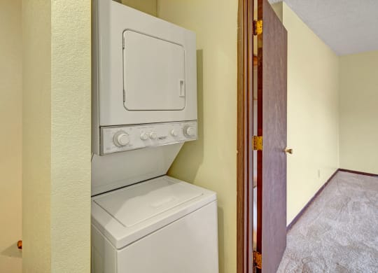 Stacked washer and dryer here at Brighton Woods Apartments in a small laundry area with beige walls. An open wooden door leads to a carpeted room, creating a cozy and tidy atmosphere.