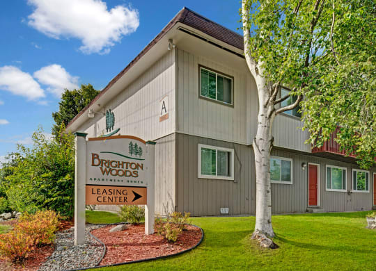 Apartment complex with a beige facade and red door, surrounded by lush greenery and birch trees. A sign reads "Brighton Woods Leasing Center."