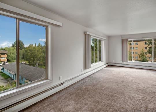 Living room here at Castle Apartments with large windows on two walls, beige carpet, and white walls. The windows offer views of trees and buildings, creating a peaceful, airy ambiance.