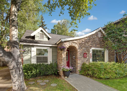 The front office here at Castle Apartments with stone facade and red door, surrounded by lush green trees and bushes. A birch tree and potted flowers add a cozy feel.