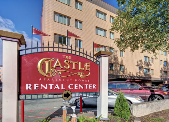 Red sign reading "The Castle Apartment Homes Rental Center" in gold and white stands prominently. Background shows a beige apartment building, parked cars, and a tree, suggesting a residential area.