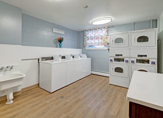 Laundry room here at Century Plaza with a cozy feeling. Features four white washers, stacked dryers, a utility sink, floral curtain, wood floor, and decorative flowers.