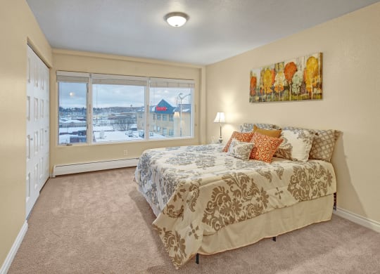A cozy bedroom here at Century Plaza with beige walls and carpet, featuring a bed with patterned bedding and colorful cushions. Large window shows a snowy view outside.