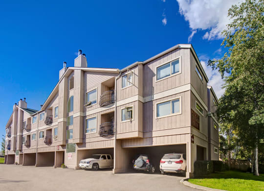 Three-story beige Chugach South Apartment building sits under a clear blue sky, with three cars parked in open garages. Trees line the right, creating a serene suburban feel.