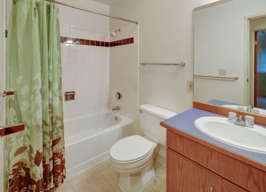 A clean, tidy bathroom here at Chugach South Apartments with a light green shower curtain featuring leaf patterns, a white bathtub, a toilet, and a wooden vanity with a blue countertop.