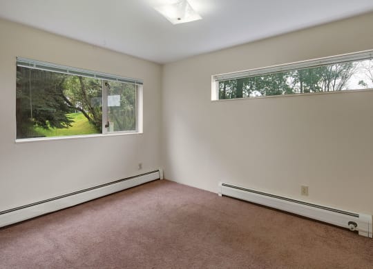 Empty living room here at Chugach South Apartements with beige carpet and cream walls, featuring two large windows showing a green, leafy backyard. The peaceful ambiance is bright and open.