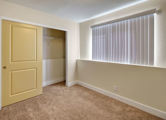 Empty bedroom here at City View Apartments with beige carpet, a partially open sliding yellow closet door on the left, and a bright window with vertical blinds on the right.
