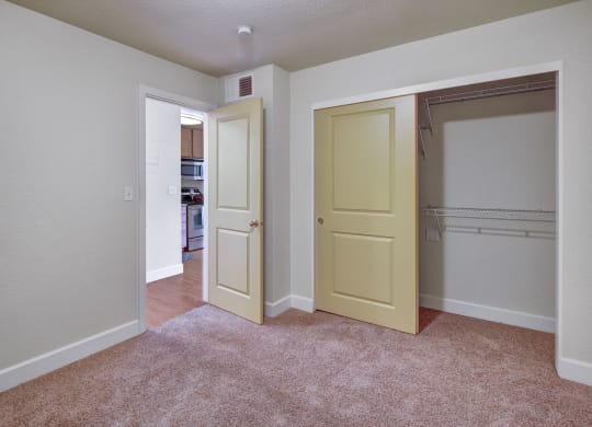 Bedroom here at City View Apartments with beige carpet and light walls. A partially open closet with sliding doors is on the right, and an open door leads to a kitchen.