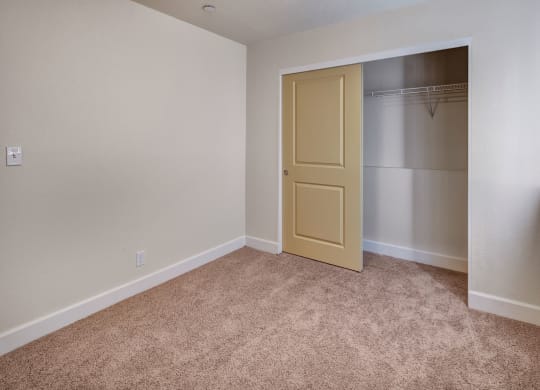 Empty beige bedroom here at City View Apartments with carpet, open closet with a sliding yellow door, white baseboards, and a small window on the right, evoking a clean, neutral feel.