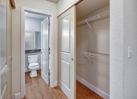 Hallway here at City View Apartments showing an open closet with wire shelves to the right, and a view into a bathroom with a white toilet and mirror on the left. Wood flooring.