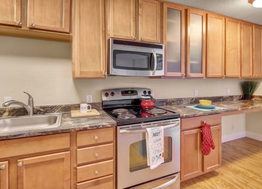 Modern kitchen here at City View Apartments with light wood cabinets, granite countertops, and stainless steel appliances. A red kettle sits on the stove, with oven mitts and towels nearby.