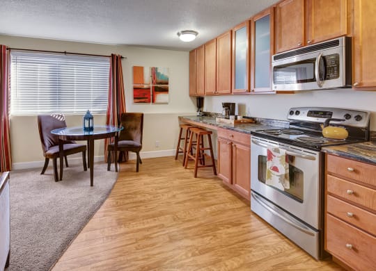 Warm kitchen-dining area here at City View Apartments with wooden cabinets, stainless steel appliances, round glass table, two chairs, large window, and abstract art on the wall.