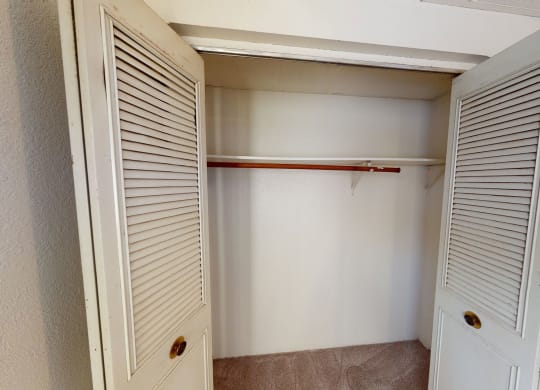 Empty closet here at College View Apartments with open louvered doors, revealing a wooden clothes rod and beige carpet. The ambiance is simple and neutral.