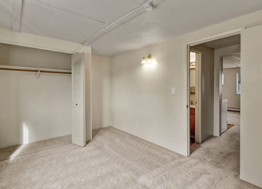 Empty beige carpeted bedroom here at College View Apartments with an open closet on the left, showing hanging rod, and an open door leading to a hallway. Dim wall light creates a cozy feel.