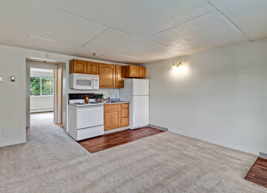Small kitchen here at College View Apartments in an apartment with wooden cabinets, white appliances, and beige carpeting. A window brightens the adjacent room, adding warmth.
