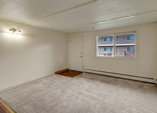 Empty living room here at College View Apartments with beige carpet, cream walls, and a white door. A window reveals an apartment building view. Overhead light casts a soft glow, creating a neutral tone.