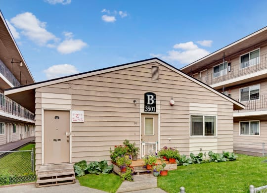 College View Apartments exterior of a beige building labeled "B 3501" with potted plants and flowers in front. Surrounding buildings have balconies. Clear blue sky above.