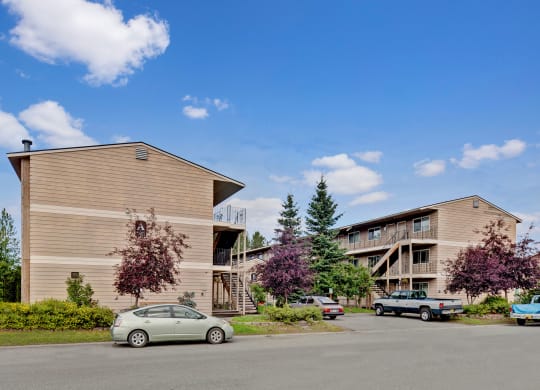 Two beige apartment buildings here at College View Apartments with wooden siding, surrounded by trees and parked cars under a clear blue sky. The scene conveys a peaceful suburban setting.