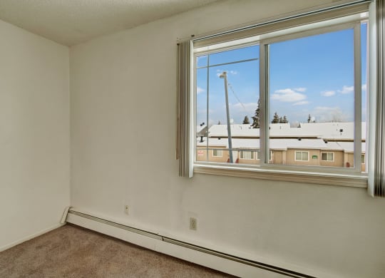 Small, empty bedroom here at Conifer Grove Apartments with beige carpet and white walls. A large window shows snowy rooftops and a clear blue sky, conveying a calm winter day.