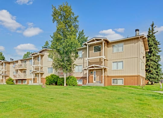 Three-story beige Conifer Grove Apartment building with white trim, surrounded by lush green grass and trees. Bright blue sky with scattered clouds, conveying a serene suburban feel.