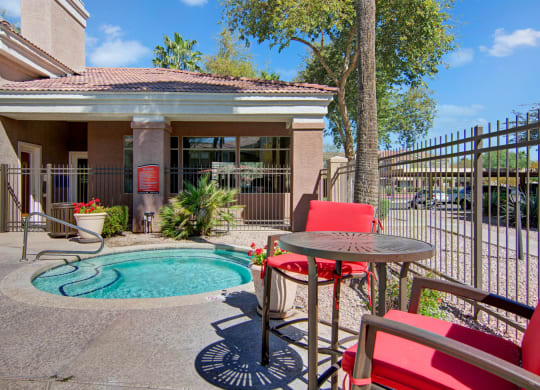 Small hot tub in a sunlit courtyard here at Dobson Towne Centre Apartments, surrounded by a brown fence and a building. Red chairs and table add a vibrant touch. Relaxing atmosphere.