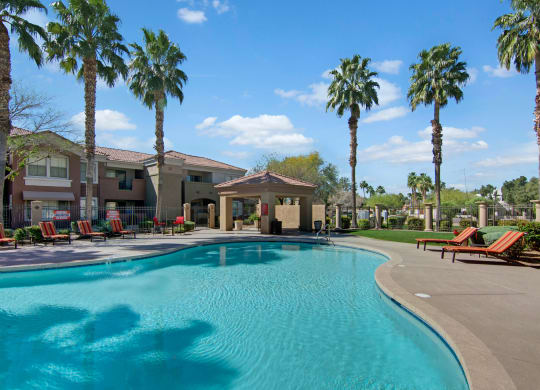 Outdoor pool area here at Dobson Towne Centre Apartments with clear blue water and red-striped lounge chairs lined up on the deck. Tall palm trees and a building are in the background under a clear blue sky.