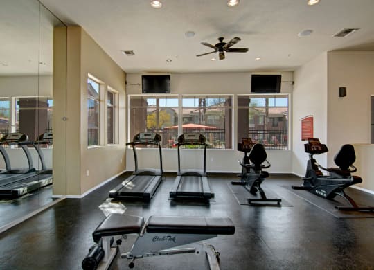 A bright gym here at Dobson Towne Centre Apartments with two treadmills and two recumbent bikes facing large windows. A weight bench is in the foreground; the atmosphere is clean and inviting.