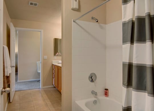 Bathroom here at Dobson Towne Centre Apartments with a white bathtub, gray-striped shower curtain, and white tiles. Beige walls lead to a hallway with a white chair visible. Cozy ambiance.