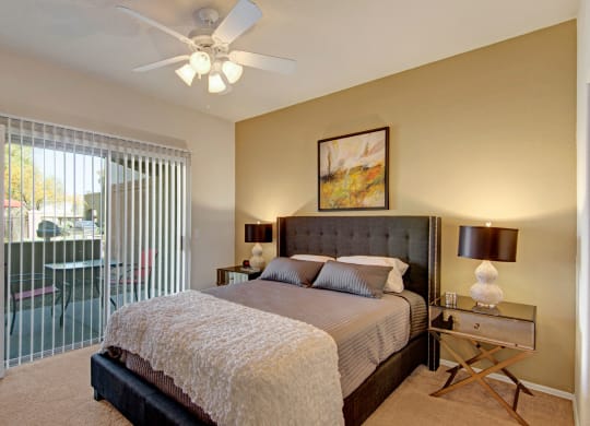 Modern bedroom here at Dobson Towne Centre Apartments with a gray upholstered bed, striped bedding, and a textured white throw. Warm lighting from side lamps, abstract art above bed, and sliding glass door with vertical blinds leading to a patio.