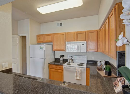 Cozy kitchen here at Dobson Towne Centre Apartments with light wood cabinets, white appliances, and a granite countertop. A kettle sits on the stove; orchids and decor add warmth.